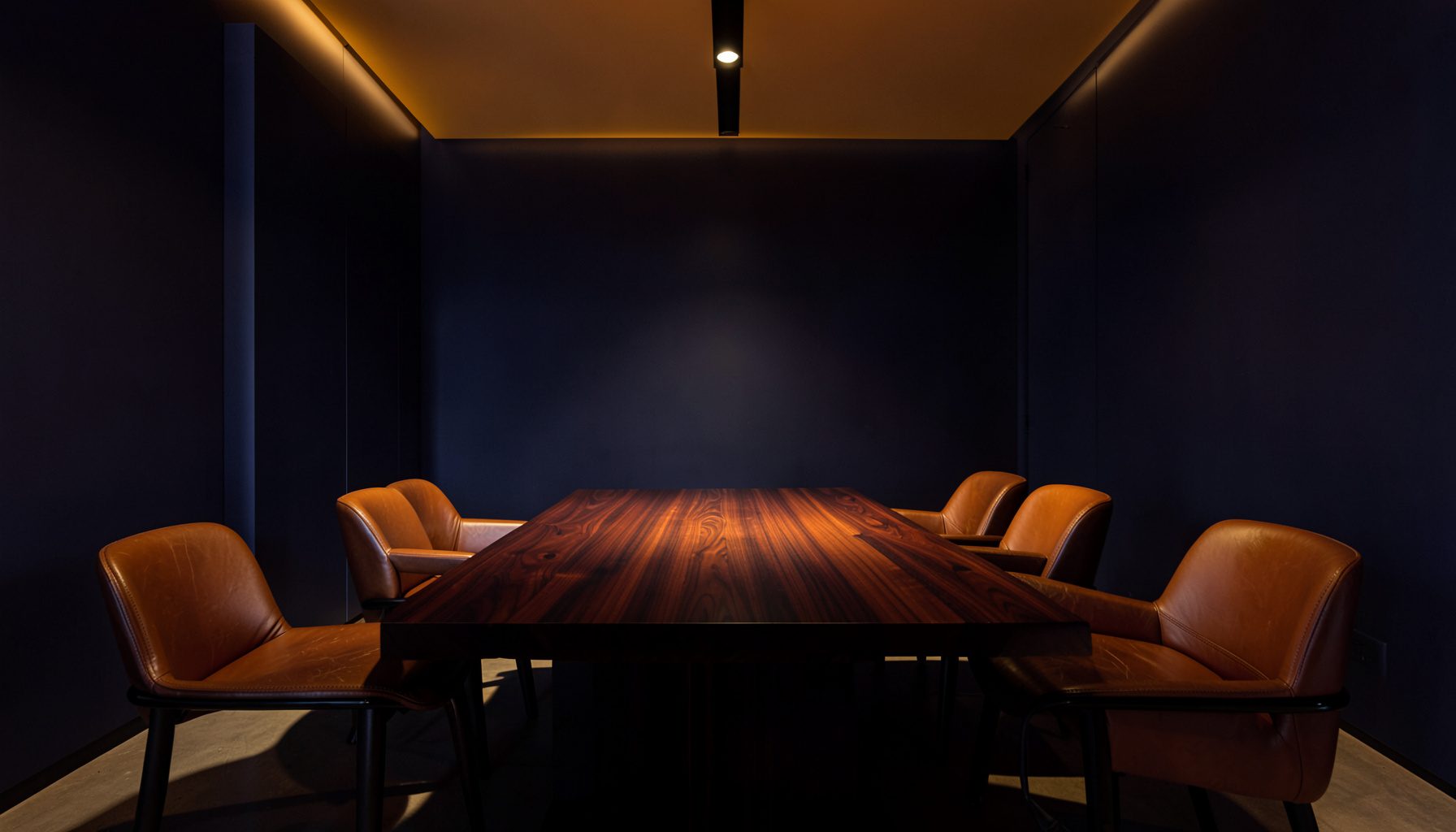 A sleek modern interior with deep navy walls, warm amber accent lighting, and a long polished dark wood table flanked by leather chairs, conveying a refined and intimate lounge atmosphere.